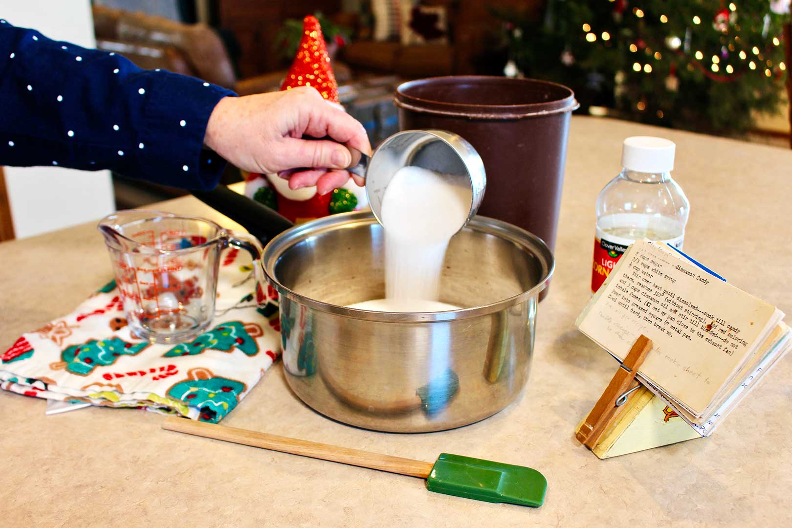 Hand pouring cup of sugar into saucepan on the countertop with other components of the cinnamon candy near by.