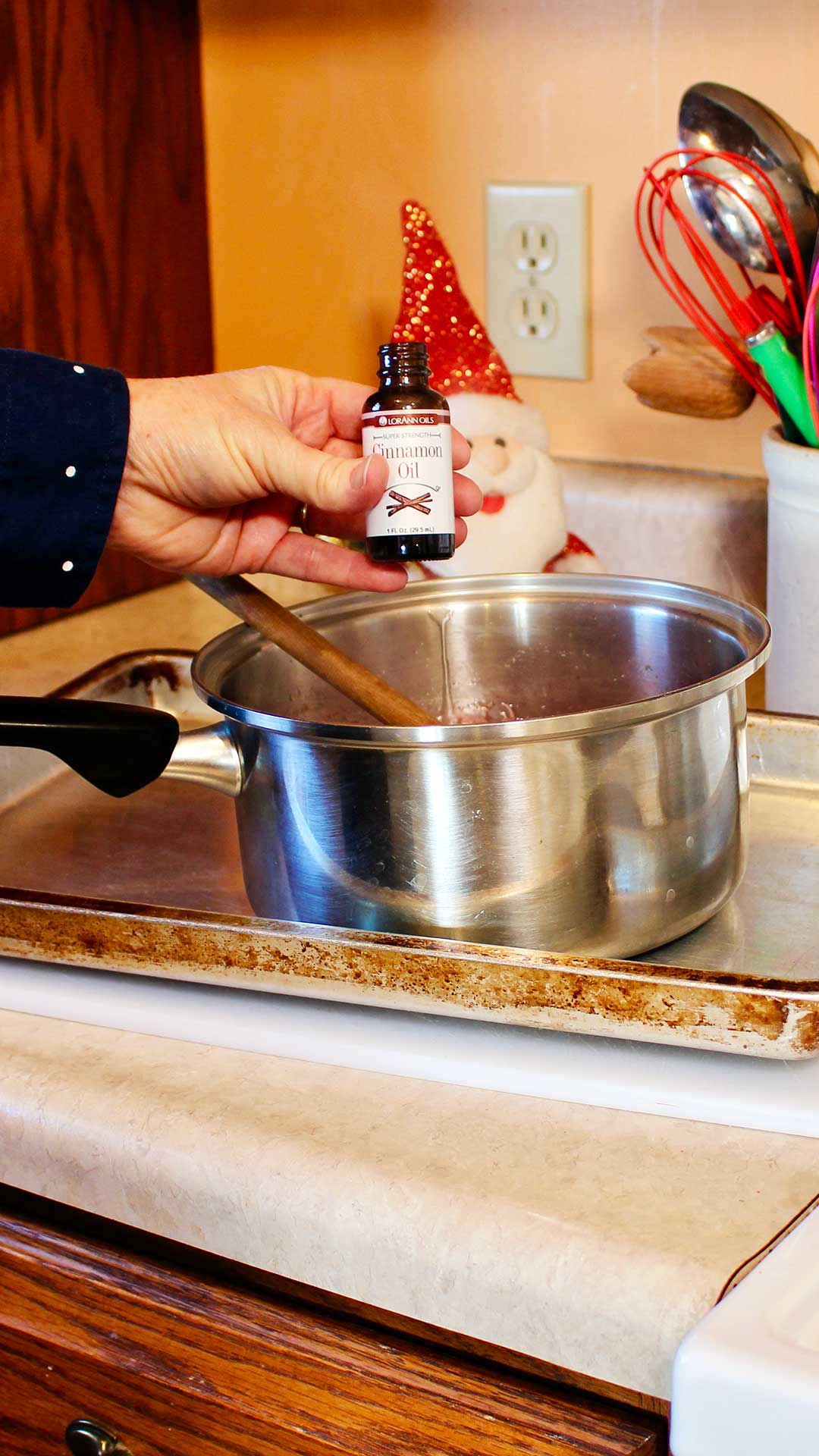 Hand holding bottle of cinnamon oil above a saucepan sitting on a cookie sheet.