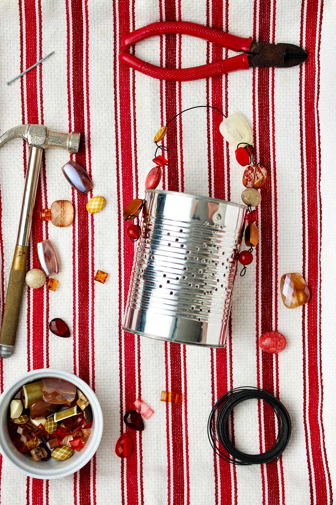 Completed Punched Tin Can Lantern resting on red and white striped linen with beads and supplies.