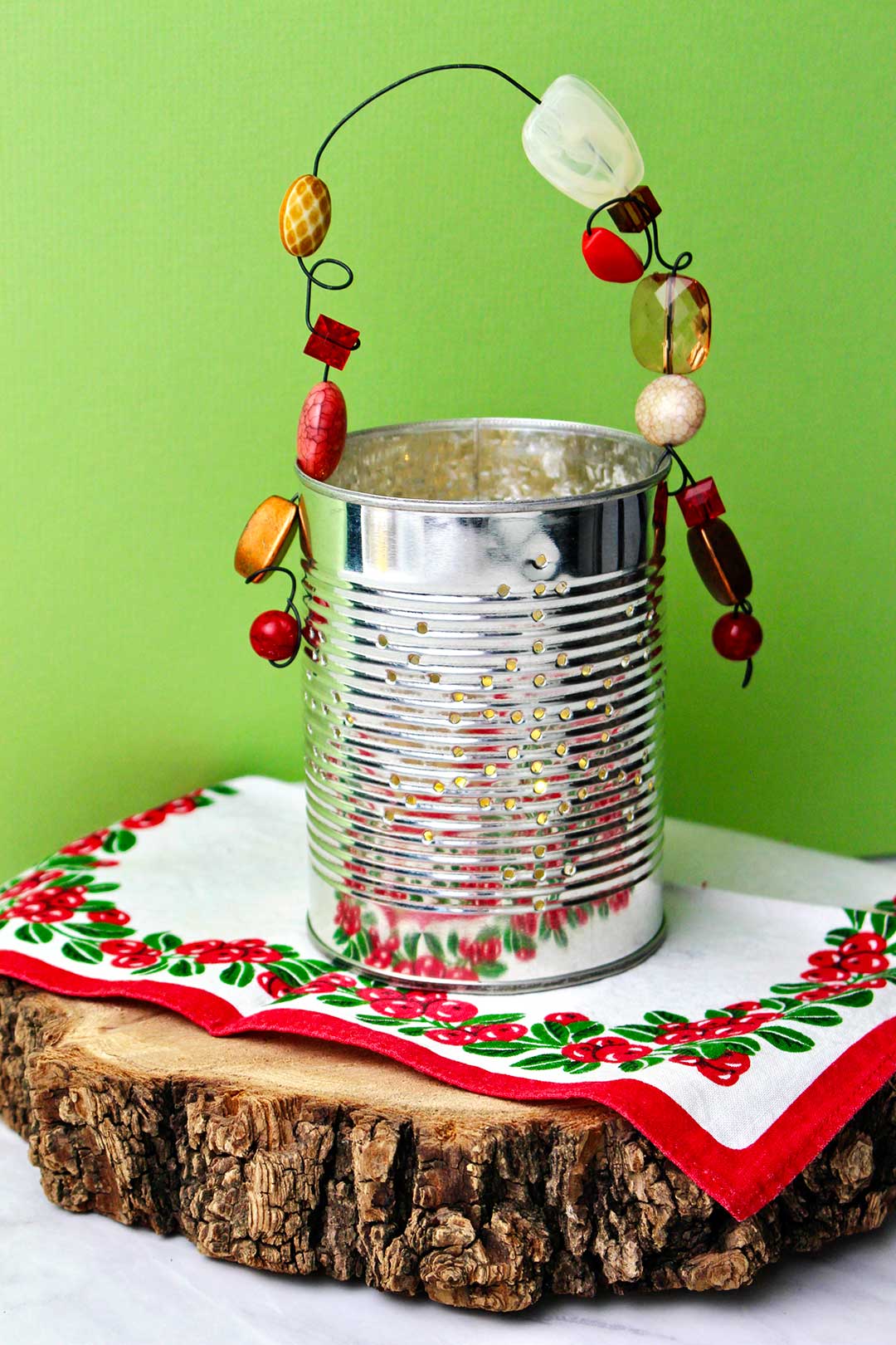 Completed Punched Tin Can Lantern resting on a slice of wood and holiday linen against a bright green background.