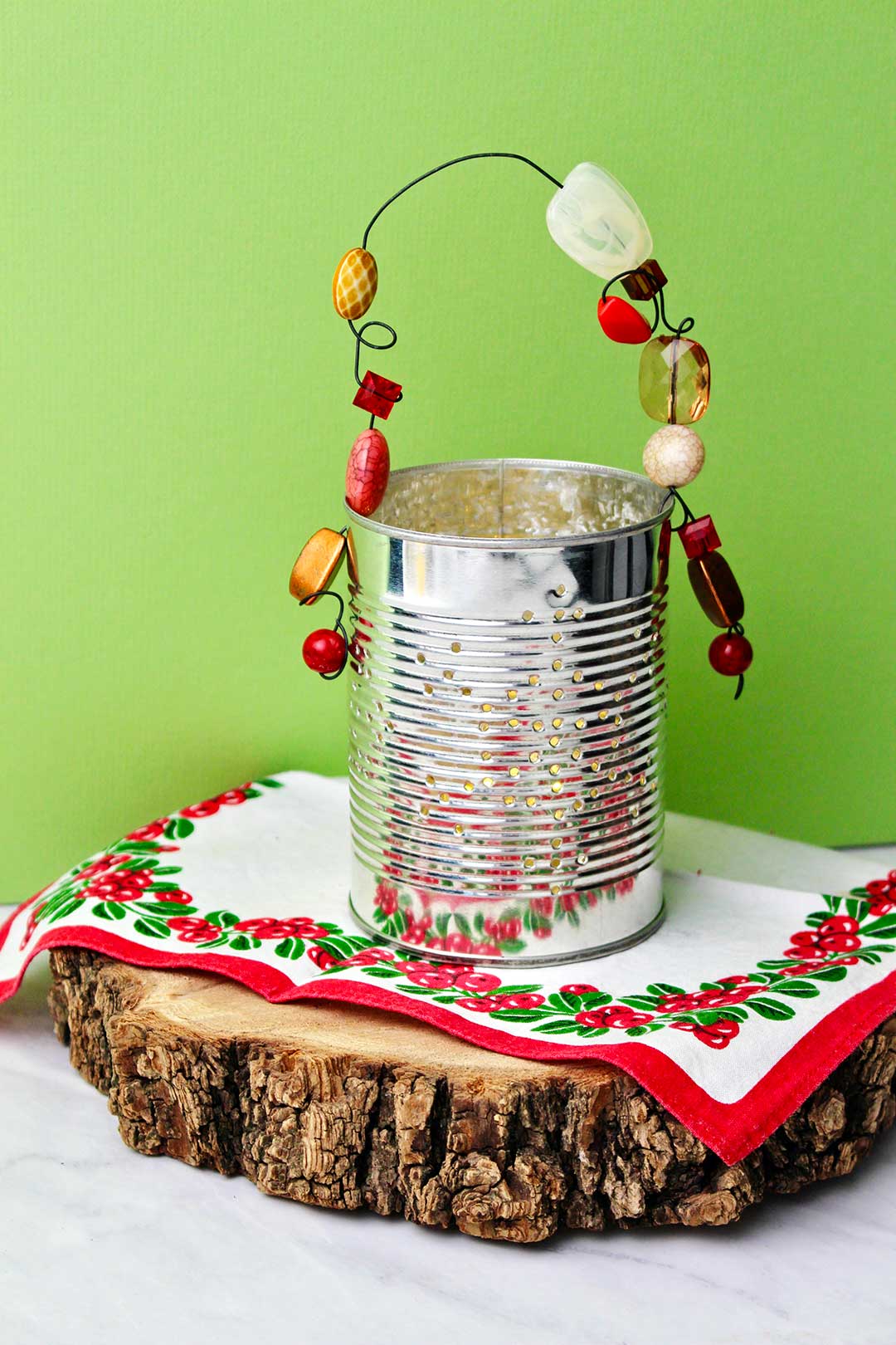 Completed Punched Tin Can Lantern resting on a slice of wood and holiday linen against a bright green background.