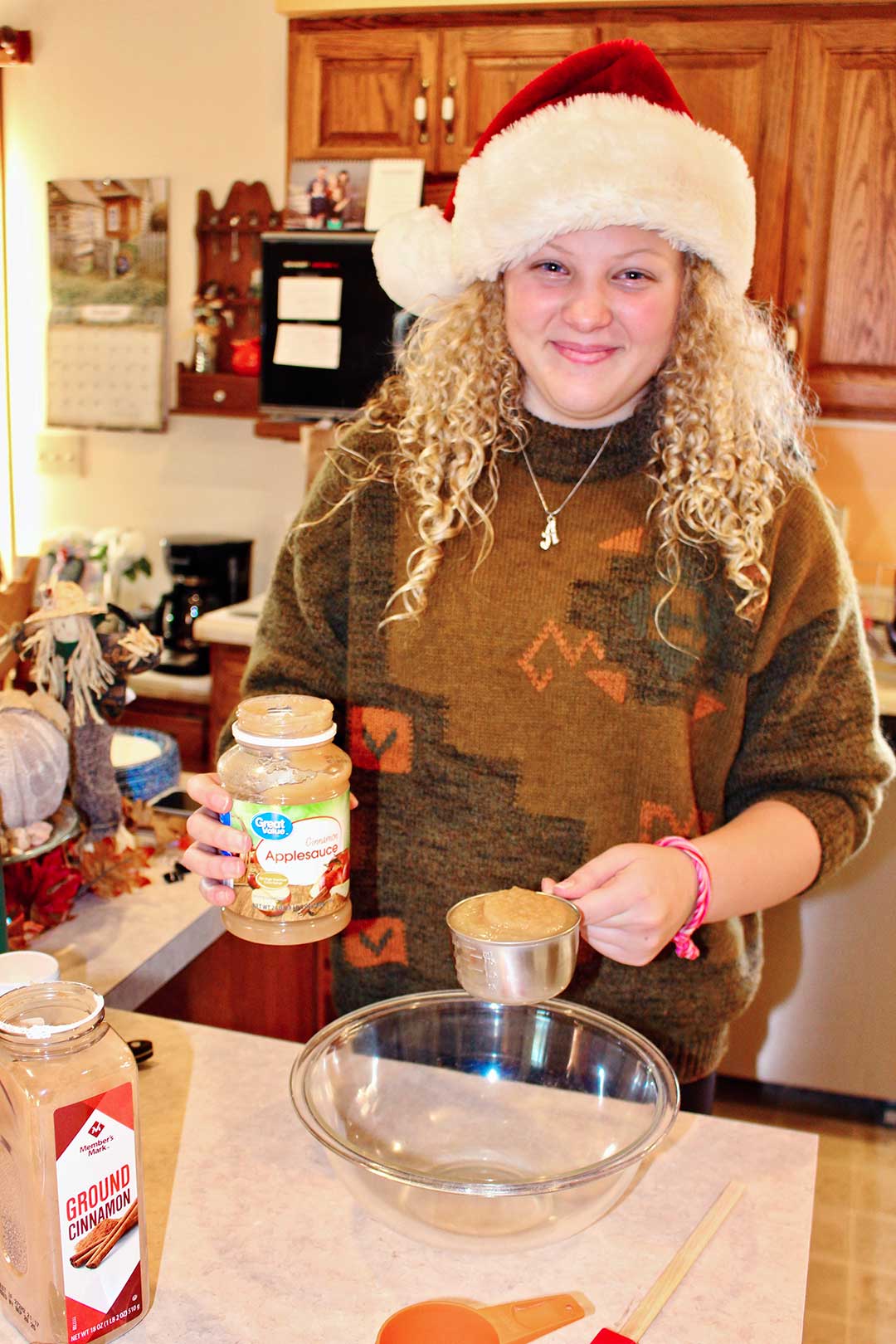 Girl with curly blonde hair and wearing a Santa hat holds a measuring cup of applesauce over a glass bowl.