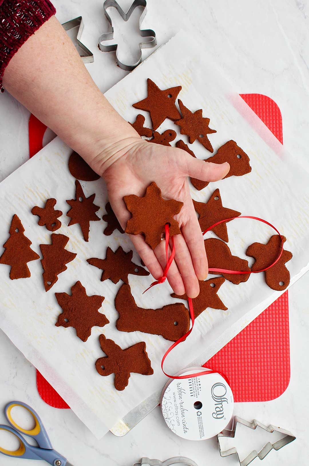 Hand holding a baked snowflake ornament with red ribbon for hanging with other ornaments on cookie sheet.