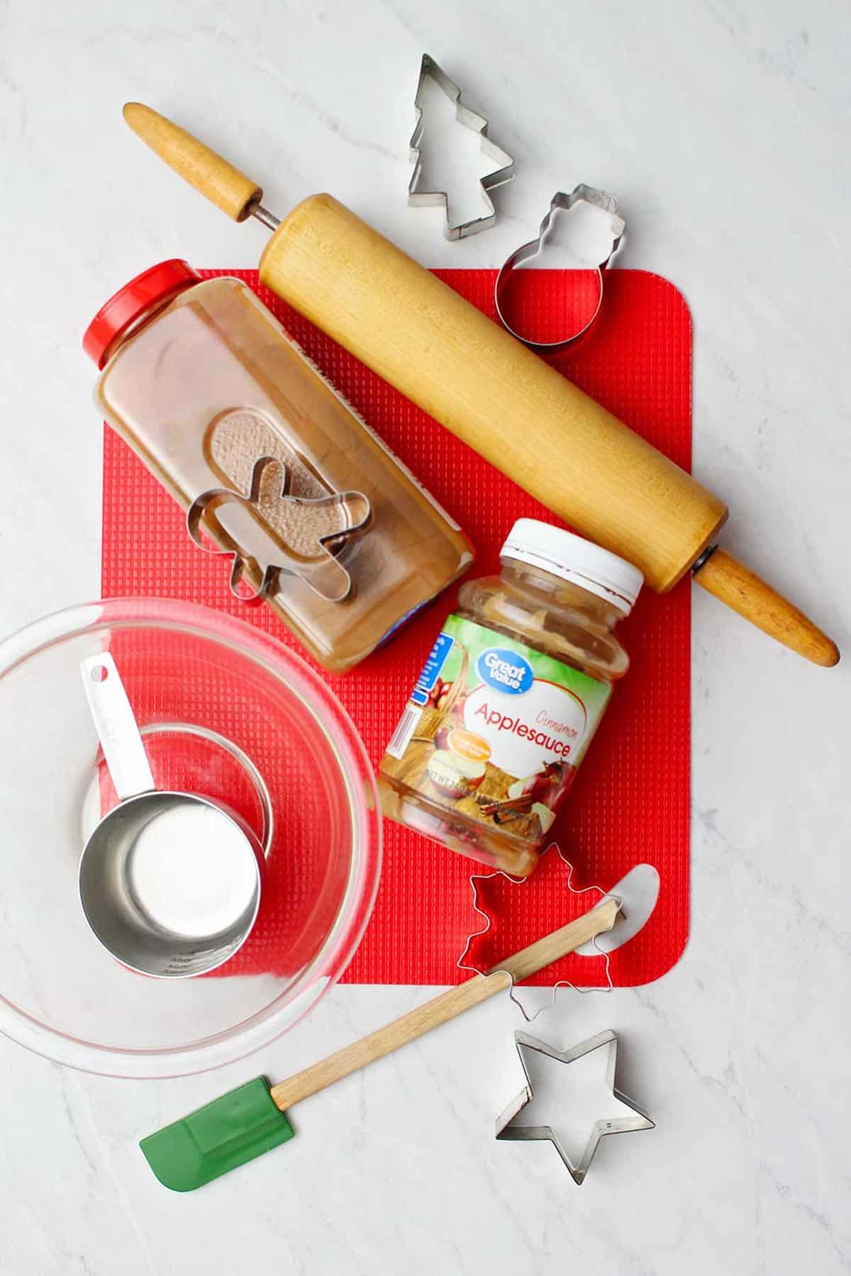 Supplies for making ornaments laying on a countertop. Cutting mat, cinnamon, applesauce, rolling pin, glass bow, and cookie cutters.