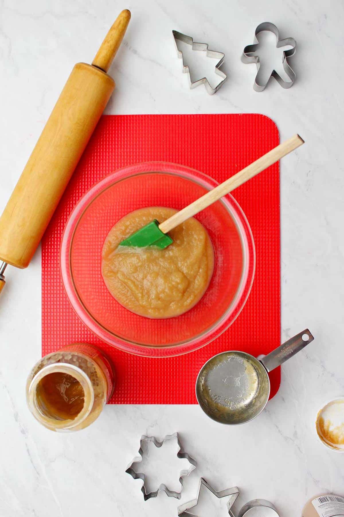 Glass bowl of applesauce with green rubber spatula resting in it sits atop a red cutting mat with various supplies around it.