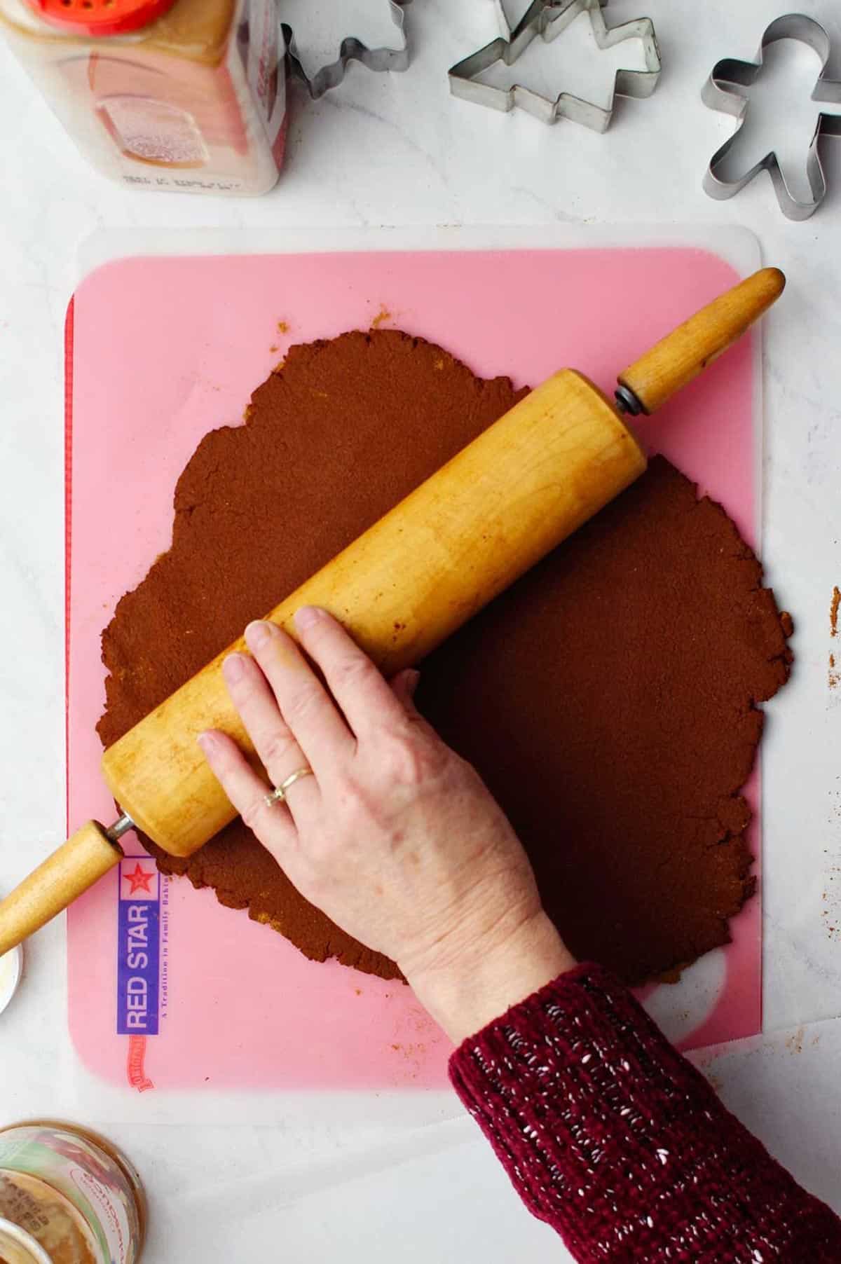 Hand rolling out the apple cinnamon dough on pink cutting mat with cookie cutters and cinnamon near by.