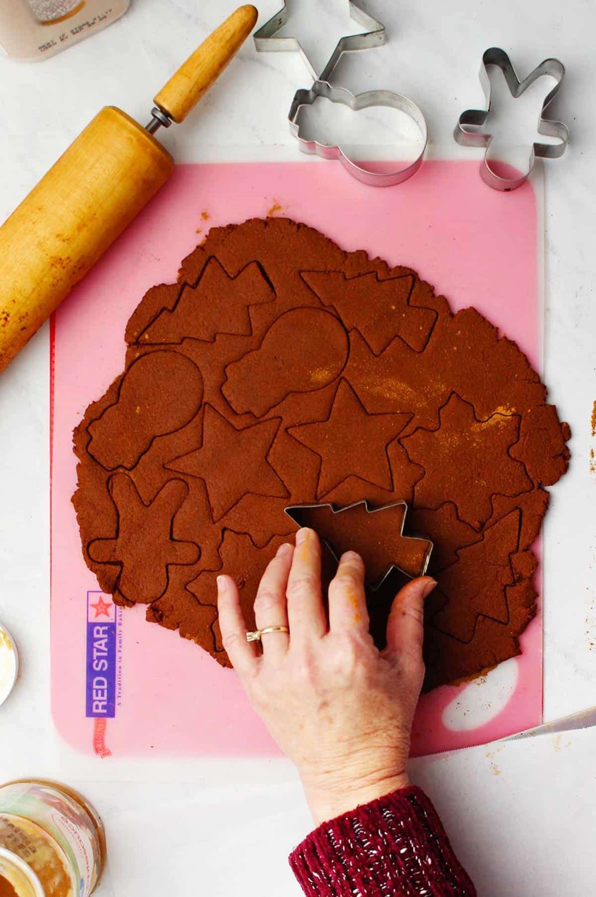 Hand pressing Christmas tree cookie cutter into rolled out dough with other shapes cut into it.