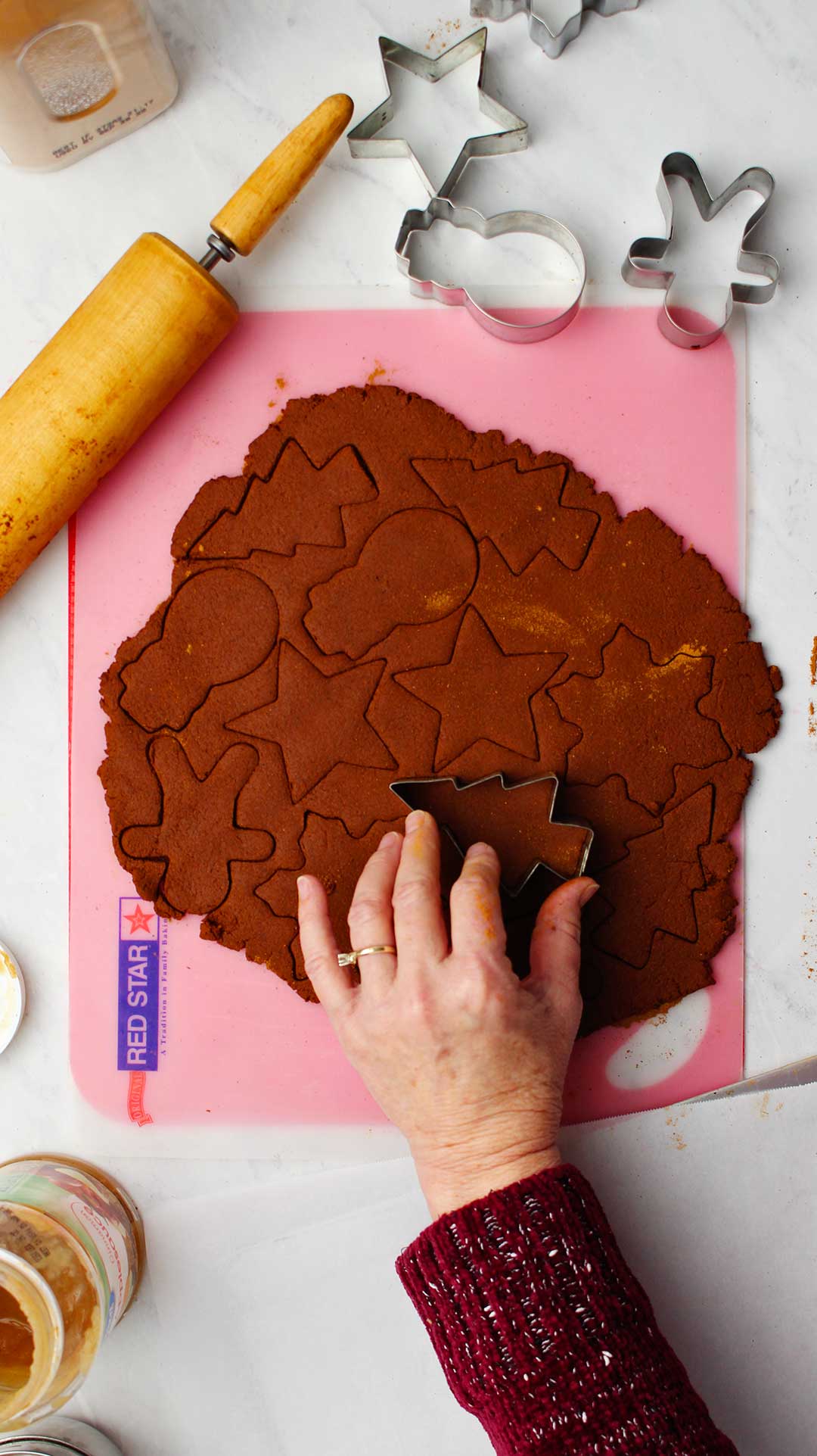 Hand pressing Christmas tree cookie cutter into rolled out dough with other shapes cut into it.