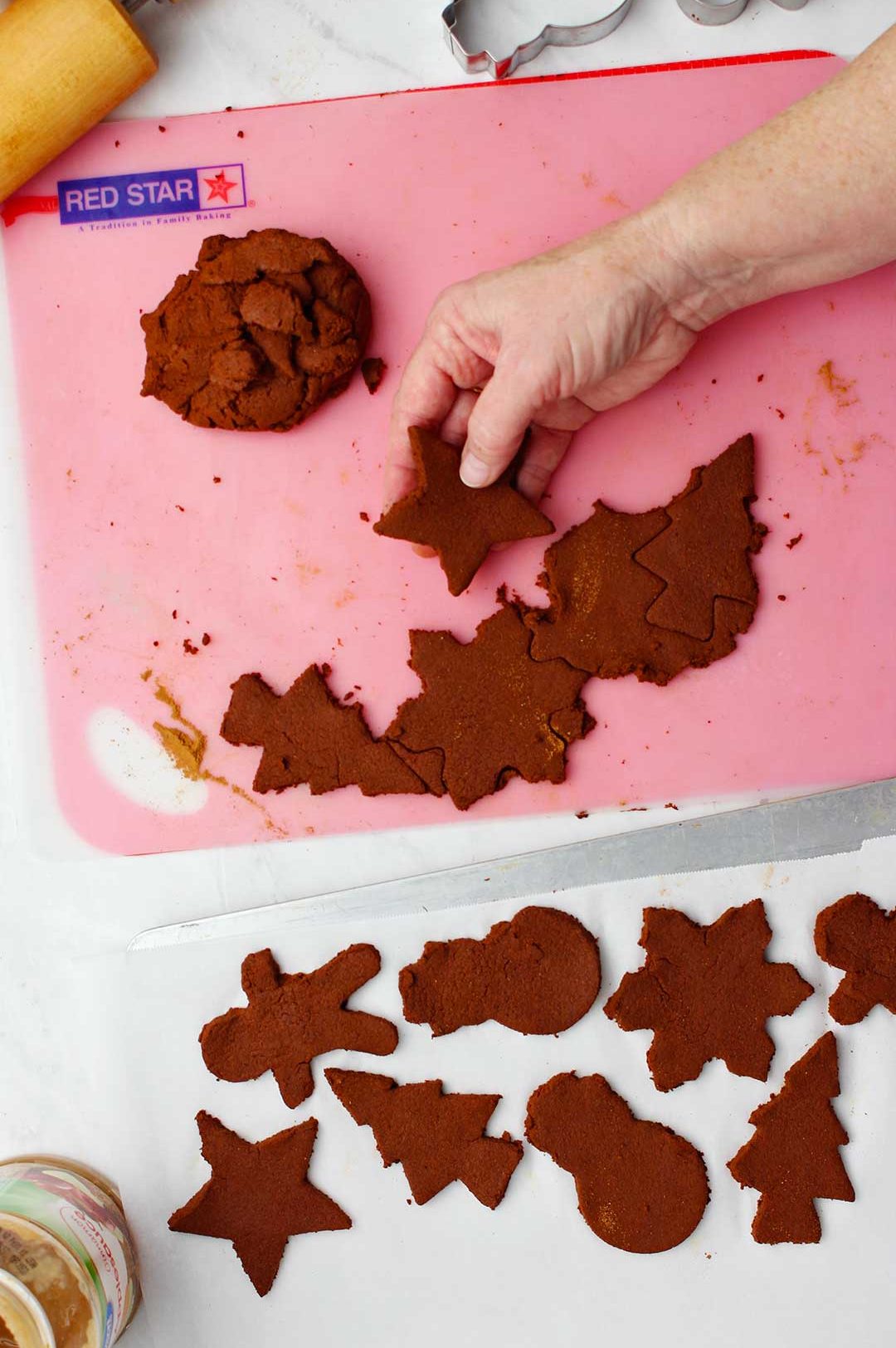 Hand removing star shaped dough from a pink cutting mat with other ornaments on parchment to the left of it.
