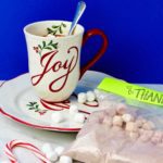 Holiday mug on saucer with marshmallows and bag of hot chocolate mix near by against a royal blue background.