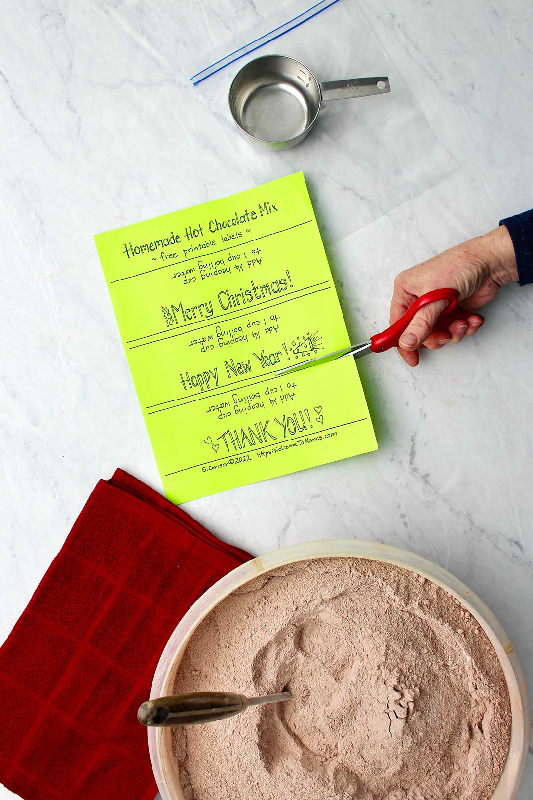 Hand cutting neon green labels for bags of hot chocolate with bowl of hot chocolate mix and red towel and cup measure near by.