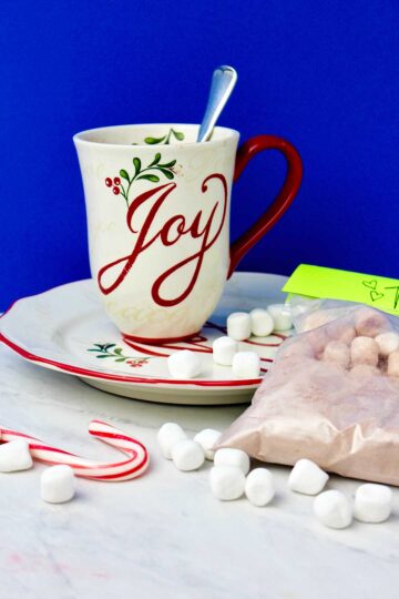 Holiday mug on saucer with marshmallows and bag of hot chocolate mix near by against a royal blue background.