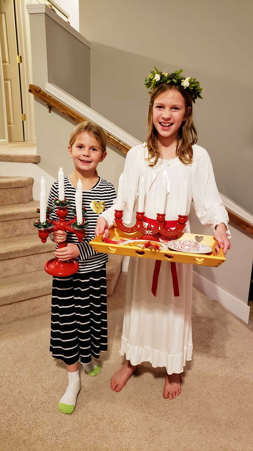 Two young girls with blonde hair, one dressed as St. Lucia, hold tray of candelabra and cookies while smiling at the camera.