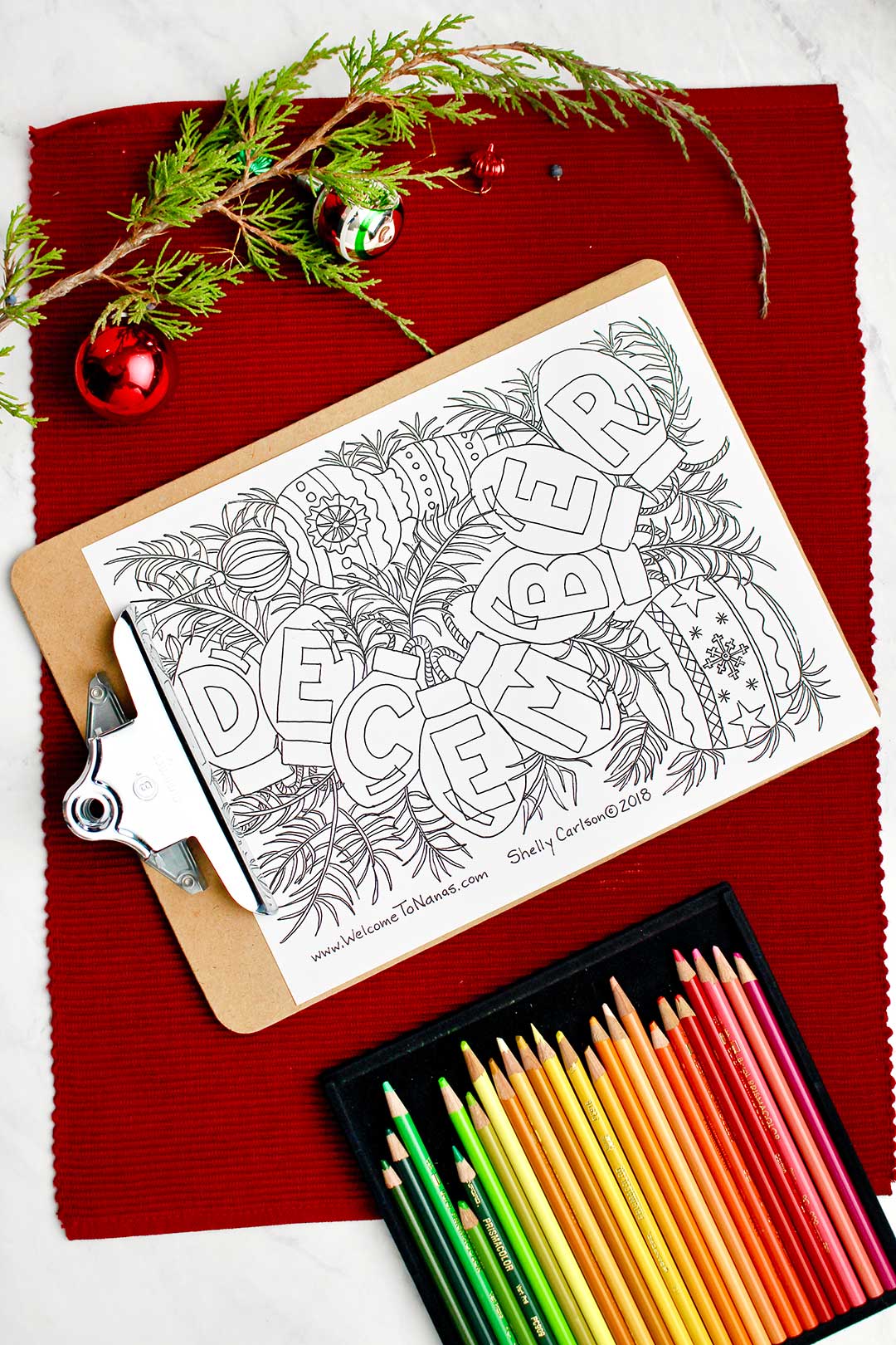 Uncolored December coloring page on a clip board resting on red placemat with holiday decorations and colored pencils near by.