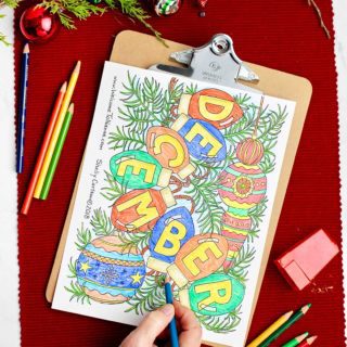 Hand coloring on the December coloring page with colored pencils and holiday decorations near by.