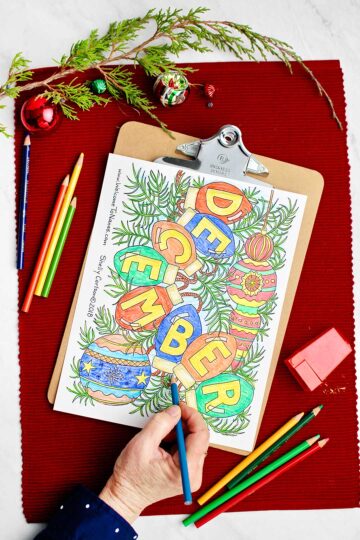 Hand coloring on the December coloring page with colored pencils and holiday decorations near by.