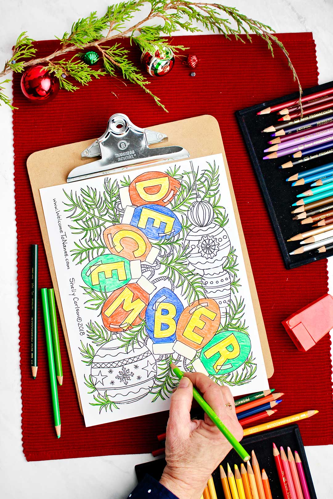 Hand coloring some greenery in the December coloring page with colored pencils and holiday decorations near by.