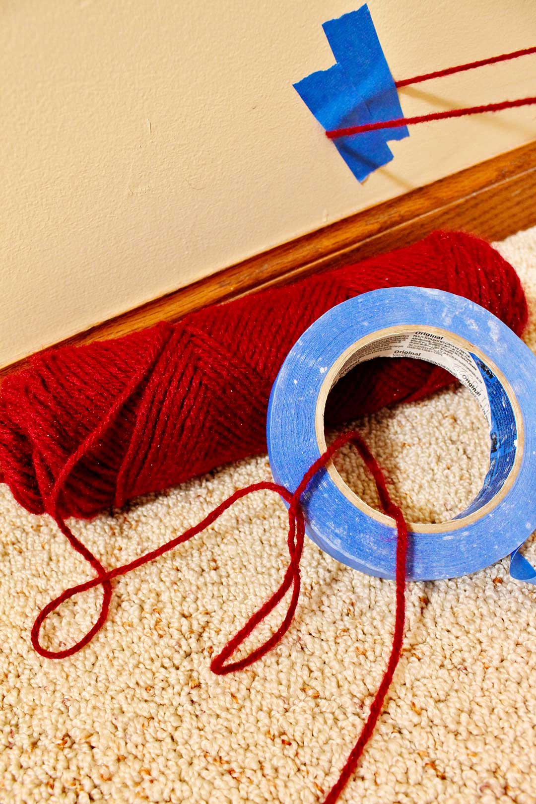 Skein of red yarn and blue painters tape lay on the carpeted floor with a piece of yarn taped to the wall with blue painters tape.