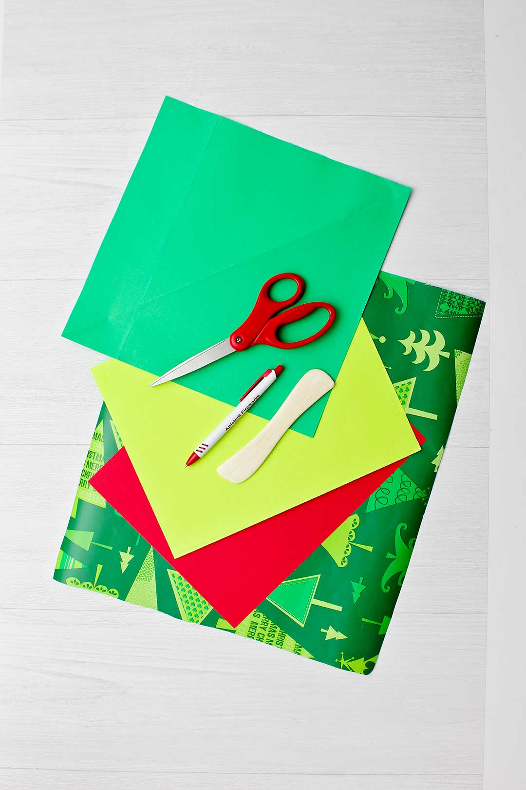 Red and green and patterned papers with scissors, pen and paper creaser laying on top.