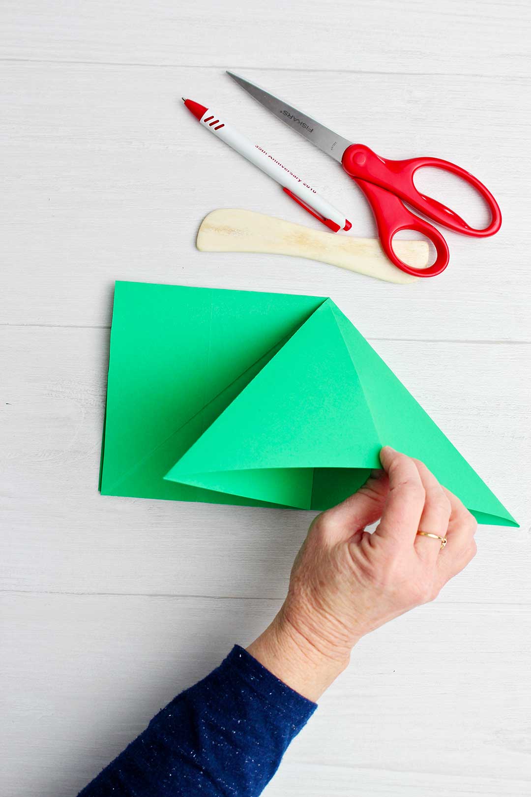 Hand holding folded piece of the green origami tree with scissors, paper creaser and pen near by.