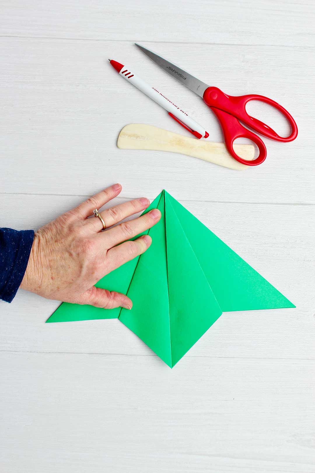 Hand holding down creased and folded pieces of the green origami tree with scissors, paper creaser and pen near by.