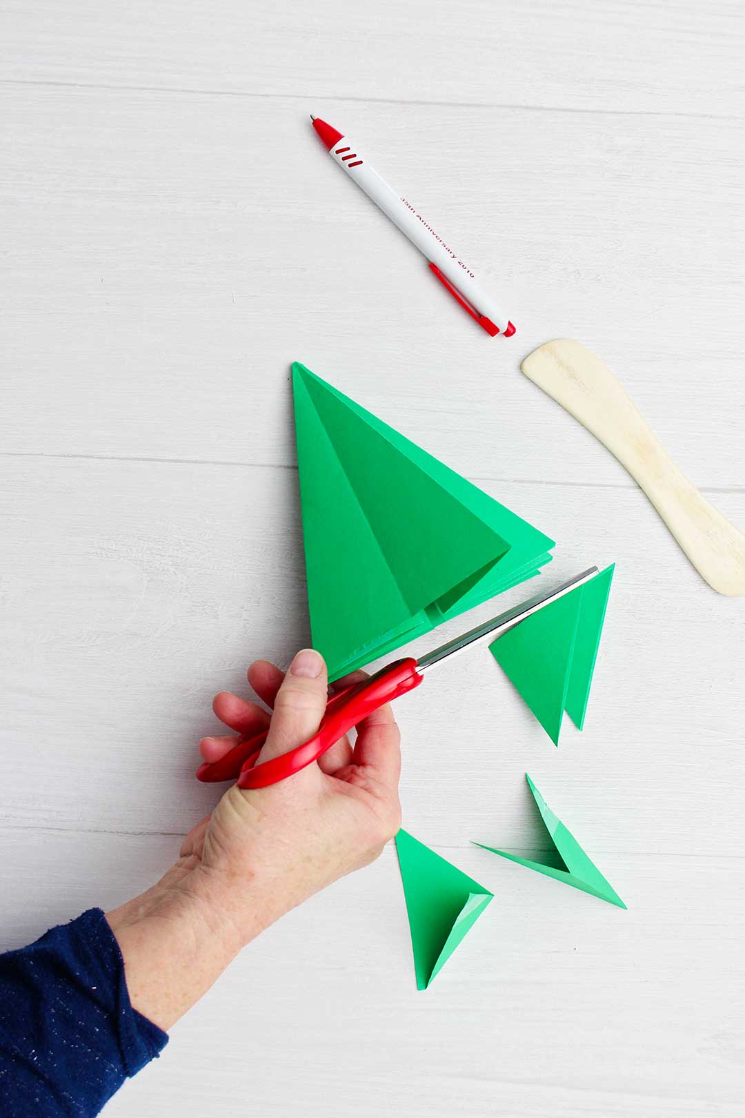 Hand trimming bottom of green origami tree with paper creaser and pen near by.