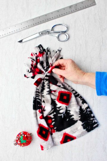 Hand holding piece of fleece that was knotted to secure pom pom on top of the hat with scissors, a ruler and pin cushion near by.