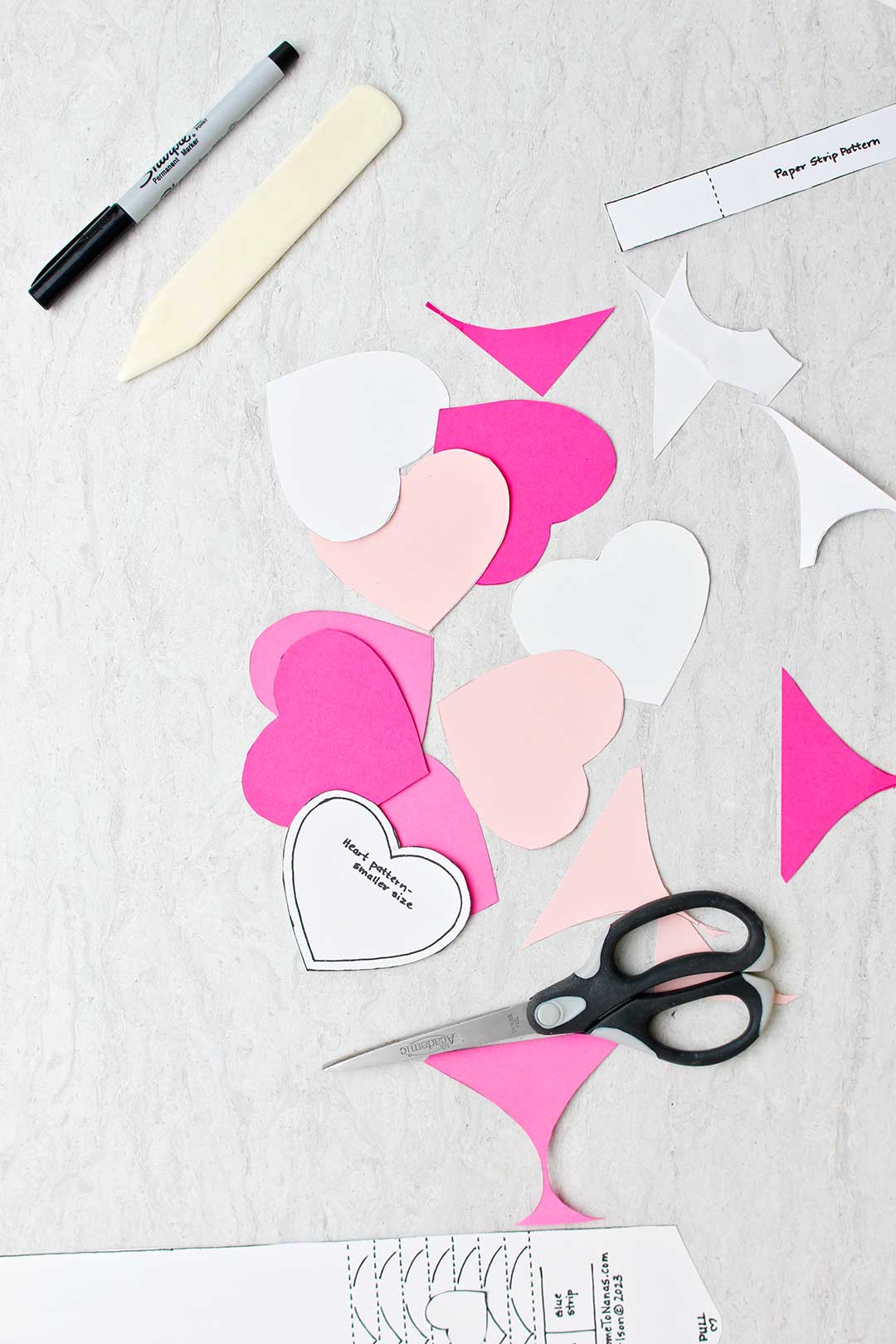 Cutouts of hearts in different shades of pink with scissors, a paper creaser, a sharpie and a template near by.