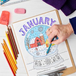 Hand shading in snow on the January coloring page on clipboard surrounded by colored pencils and purple gloves.
