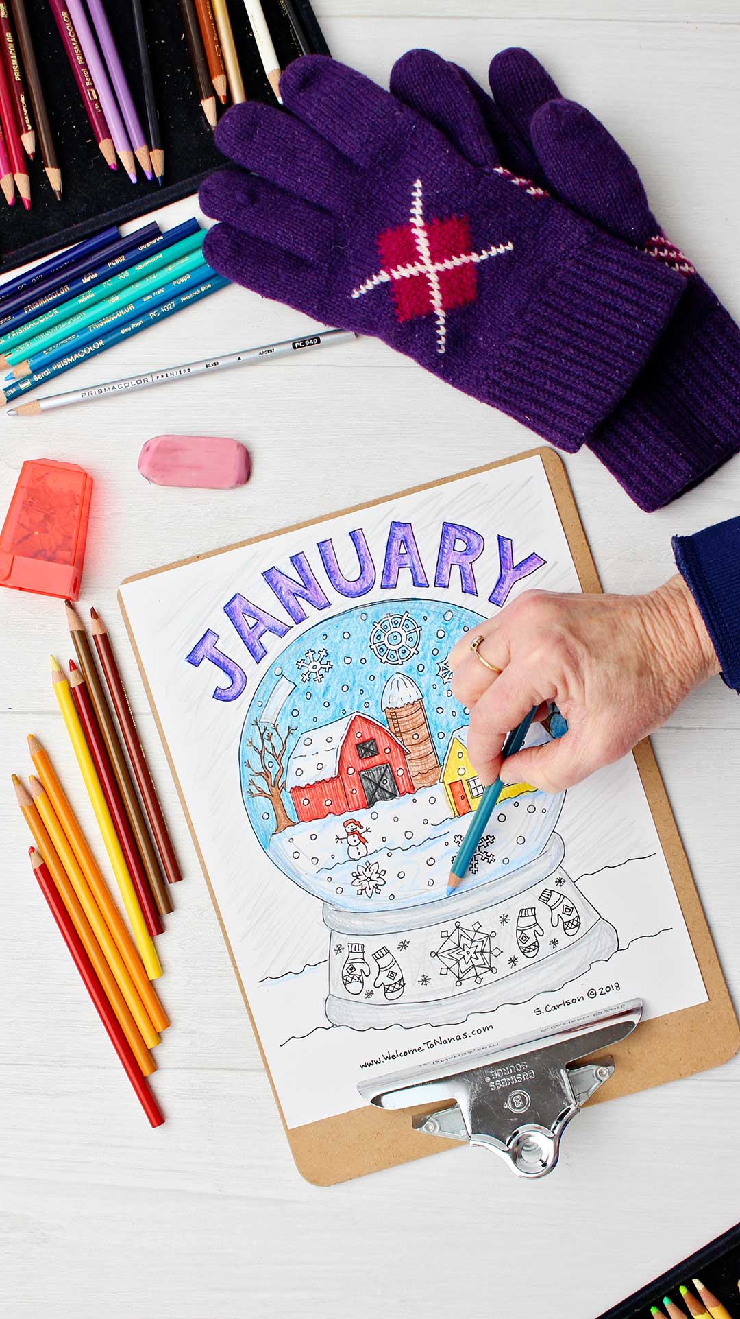 Hand shading in snow on the January coloring page on clipboard surrounded by colored pencils and purple gloves.