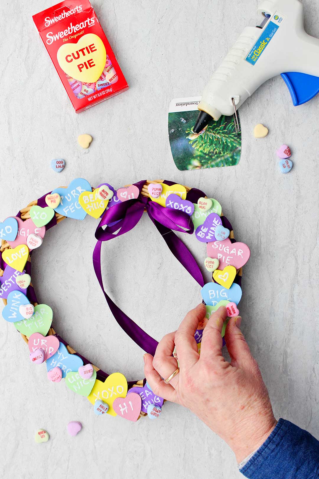 Hand placing candy conversation heart on wreath to fill in some spaces in between larger wooden conversation hearts.