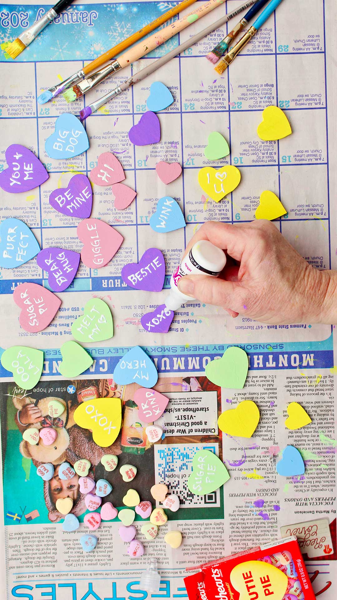 Hand using white puff paint to write messages on wooden conversation hearts.