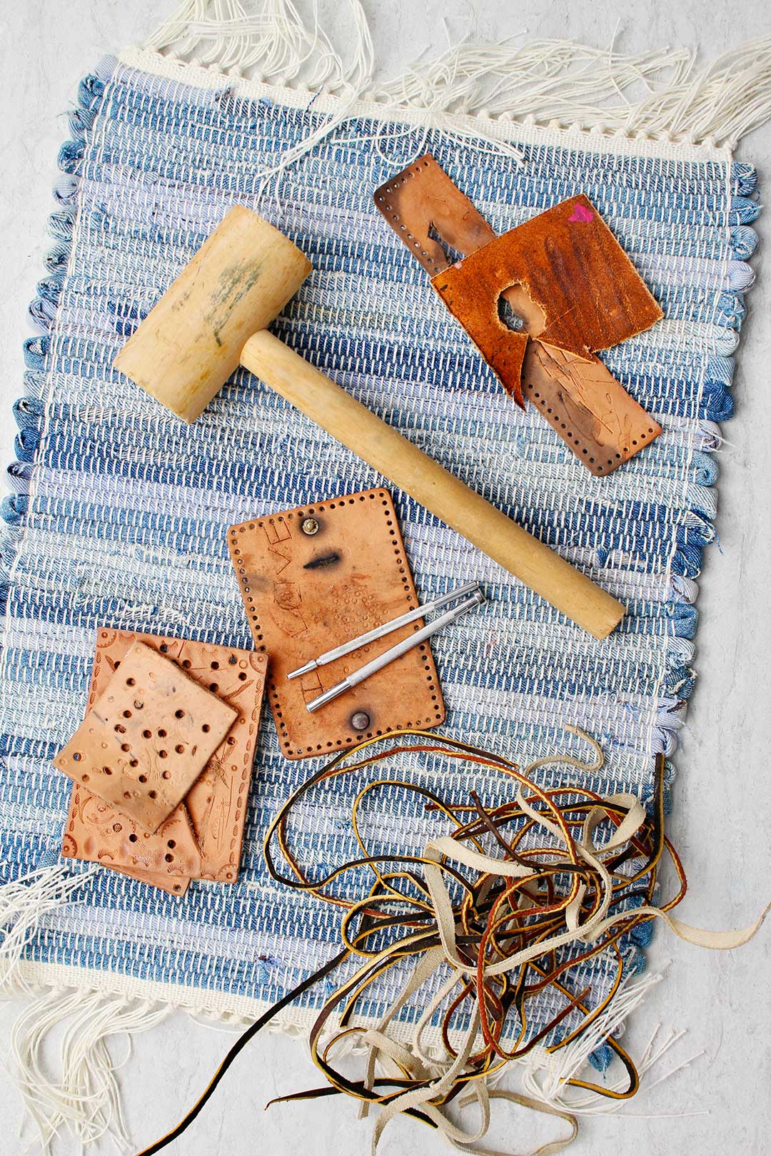 Supplies for making Leather Tassel Keychains on a blue placemat. Wooden mallet, leather pieces and puncturing tools.