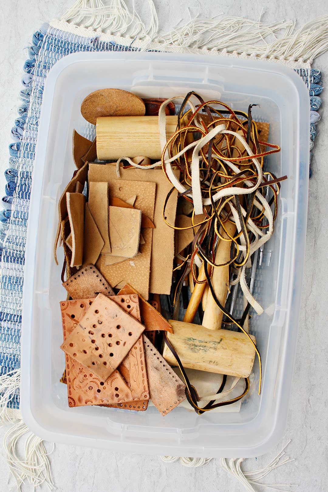 A clear Tupperware full of leather pieces and wooden mallets sitting on a blue placemat.