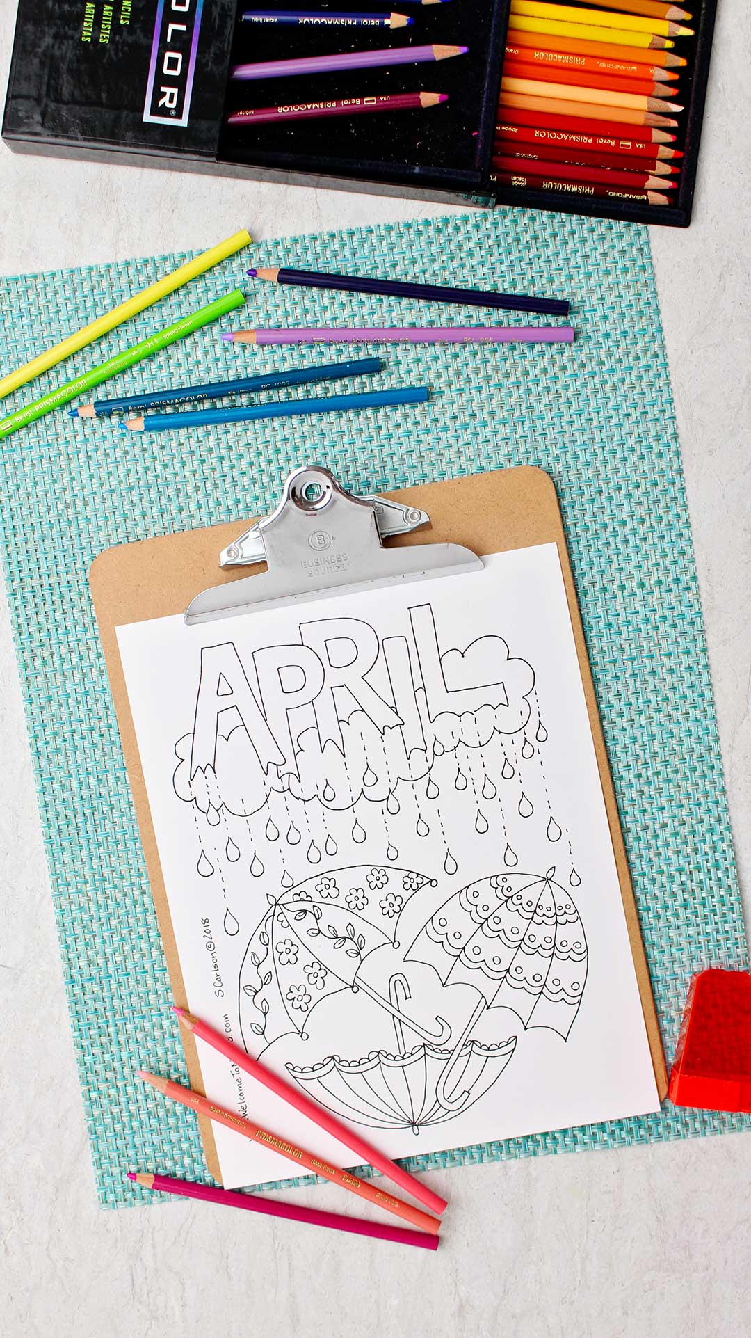 Uncolored April Showers Coloring Page on clipboard resting on an aqua placemat with colored pencils around.
