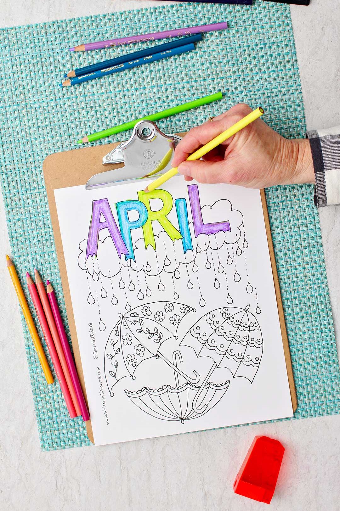 Hand coloring the "R" in April bright green in the April Showers Coloring Page sitting on an aqua placemat with colored pencils around.