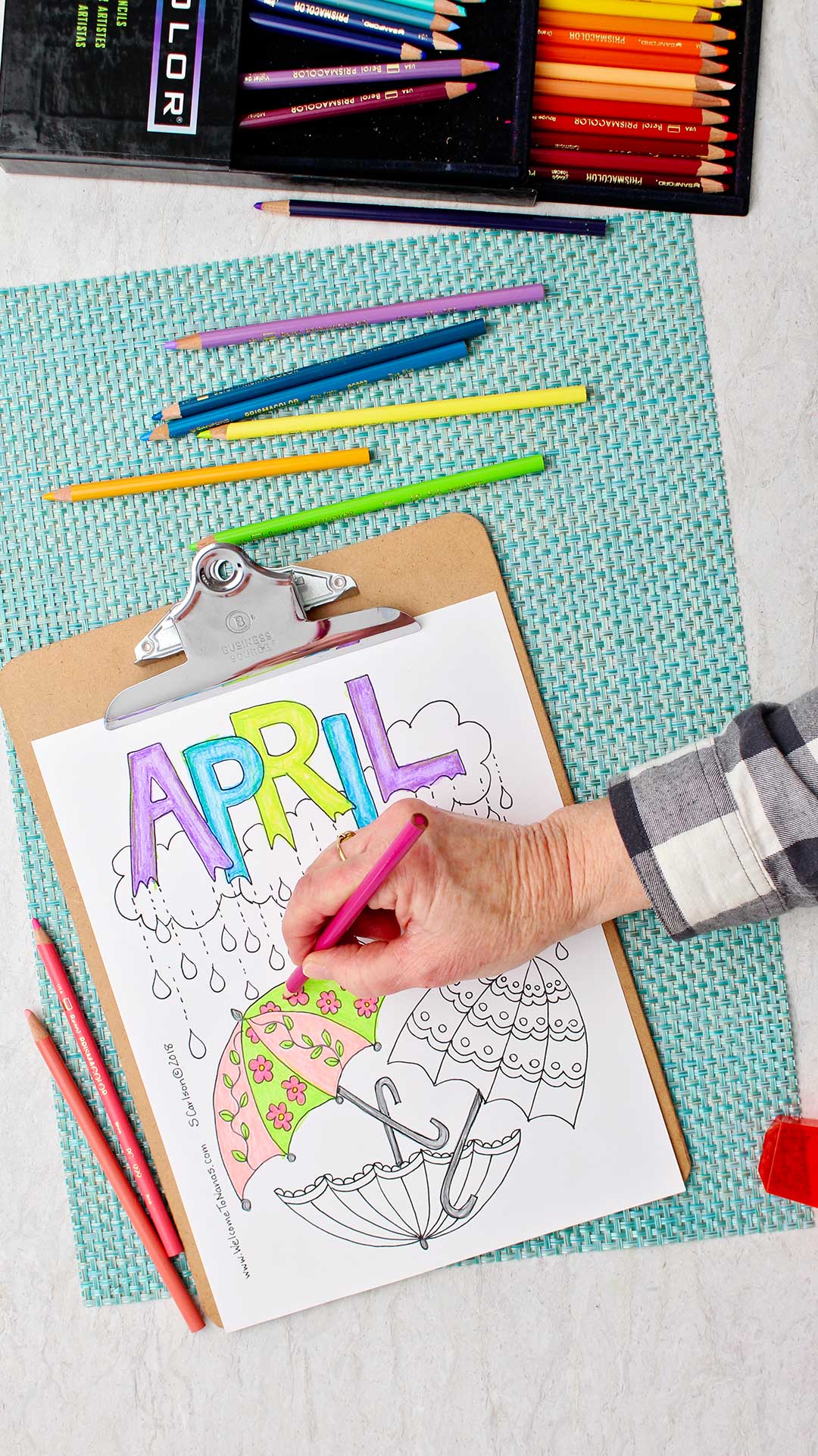 Hand coloring flowers on an umbrella pink in the April Showers Coloring Page sitting on an aqua placemat with colored pencils around.
