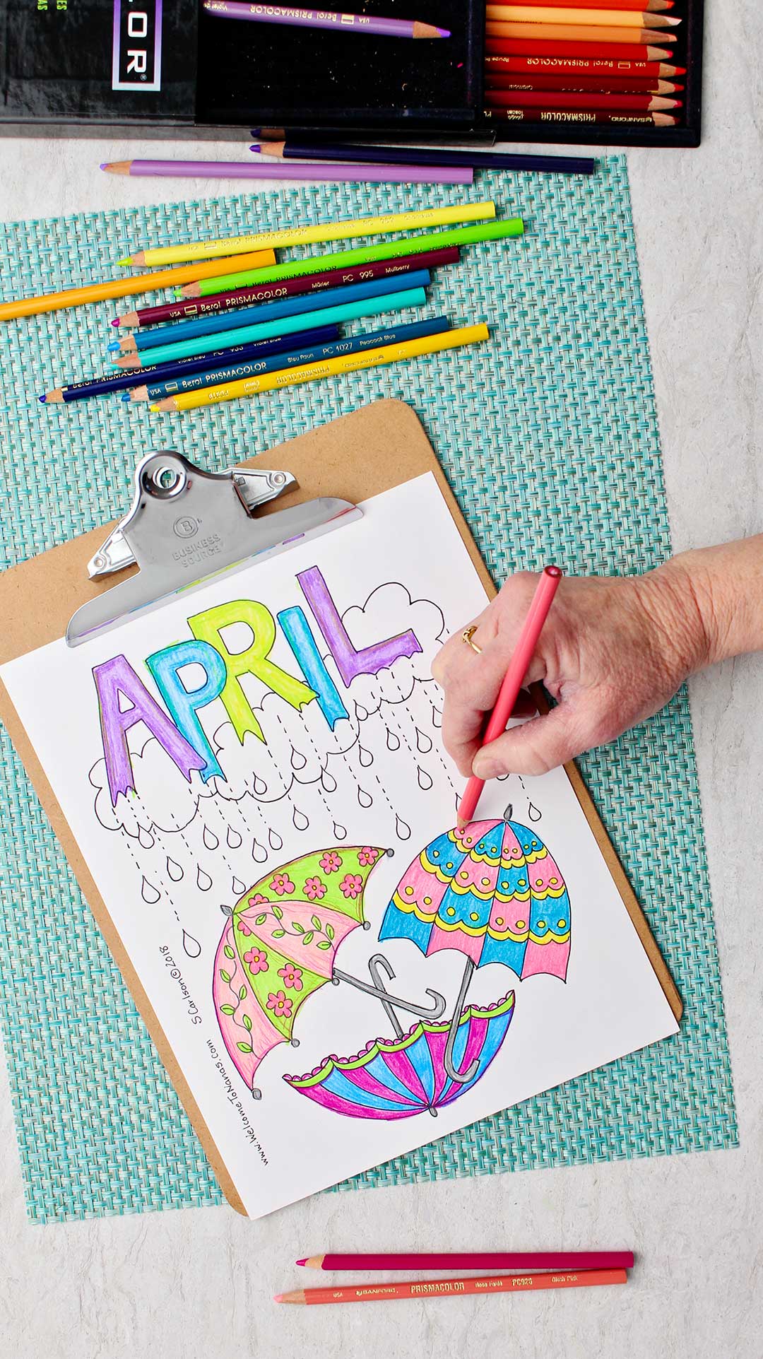 Hand coloring part of an umbrella pink in the April Showers Coloring Page sitting on an aqua placemat with colored pencils around.