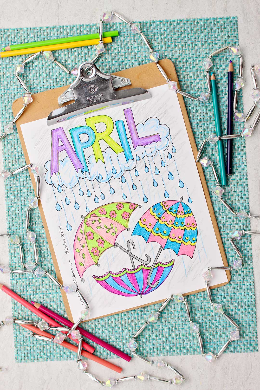 Completed April coloring page colored in shades of blue, pink, purple and green, sitting on an aqua placemat surrounded by a string of clear and silver beads and colored pencils.