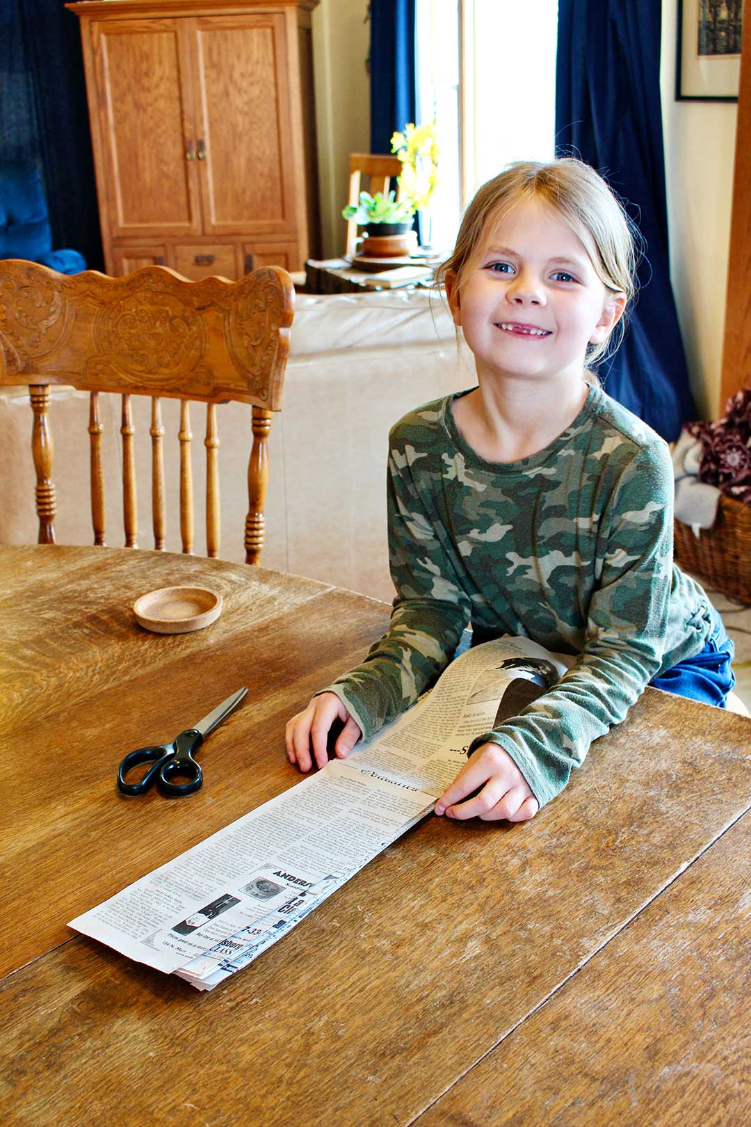 Young girl with blonde hair, missing front teeth and a camp shirt smiles at camera while at kitchen table holding large strips of newspaper.