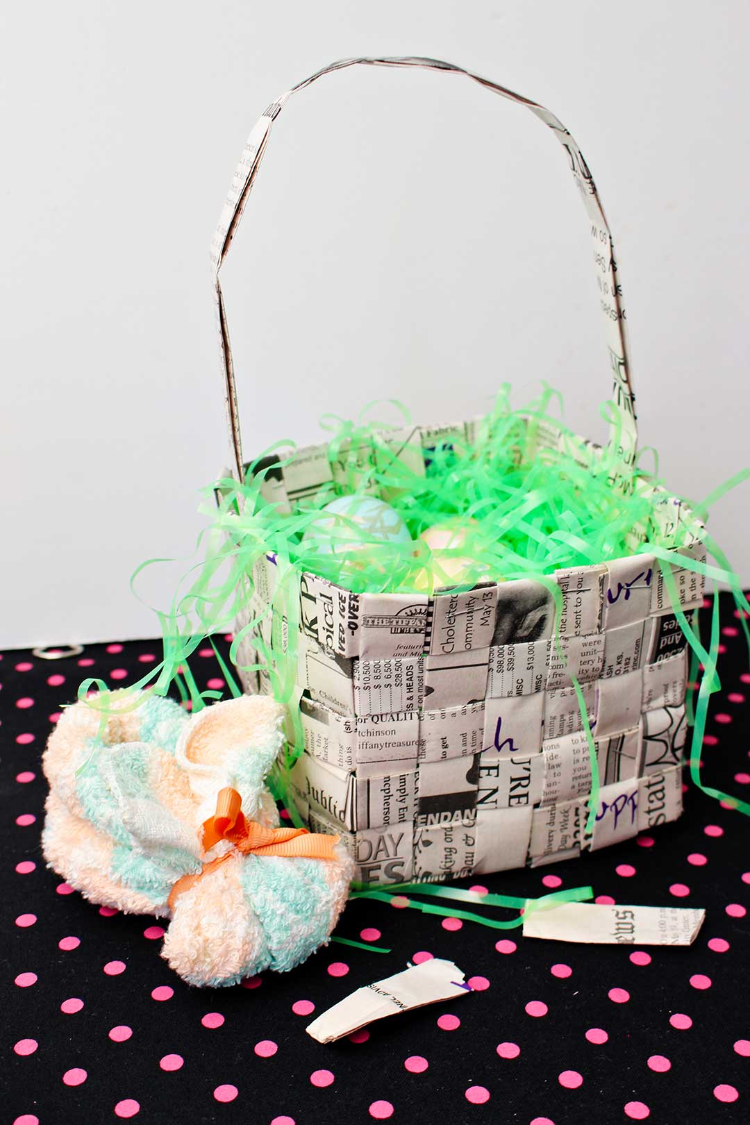 Completed woven newspaper basket filled with easter basket grass and eggs resting on pink polka dotted black fabric.