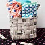 Completed woven newspaper basket full of colorful hand towels sitting on a piece of pink polka dotted black fabric.