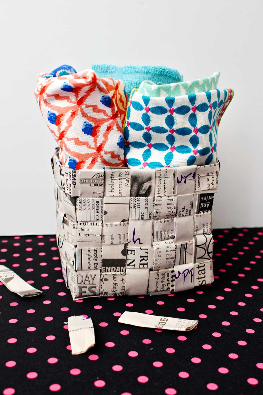 Completed woven newspaper basket full of colorful hand towels sitting on a piece of pink polka dotted black fabric.