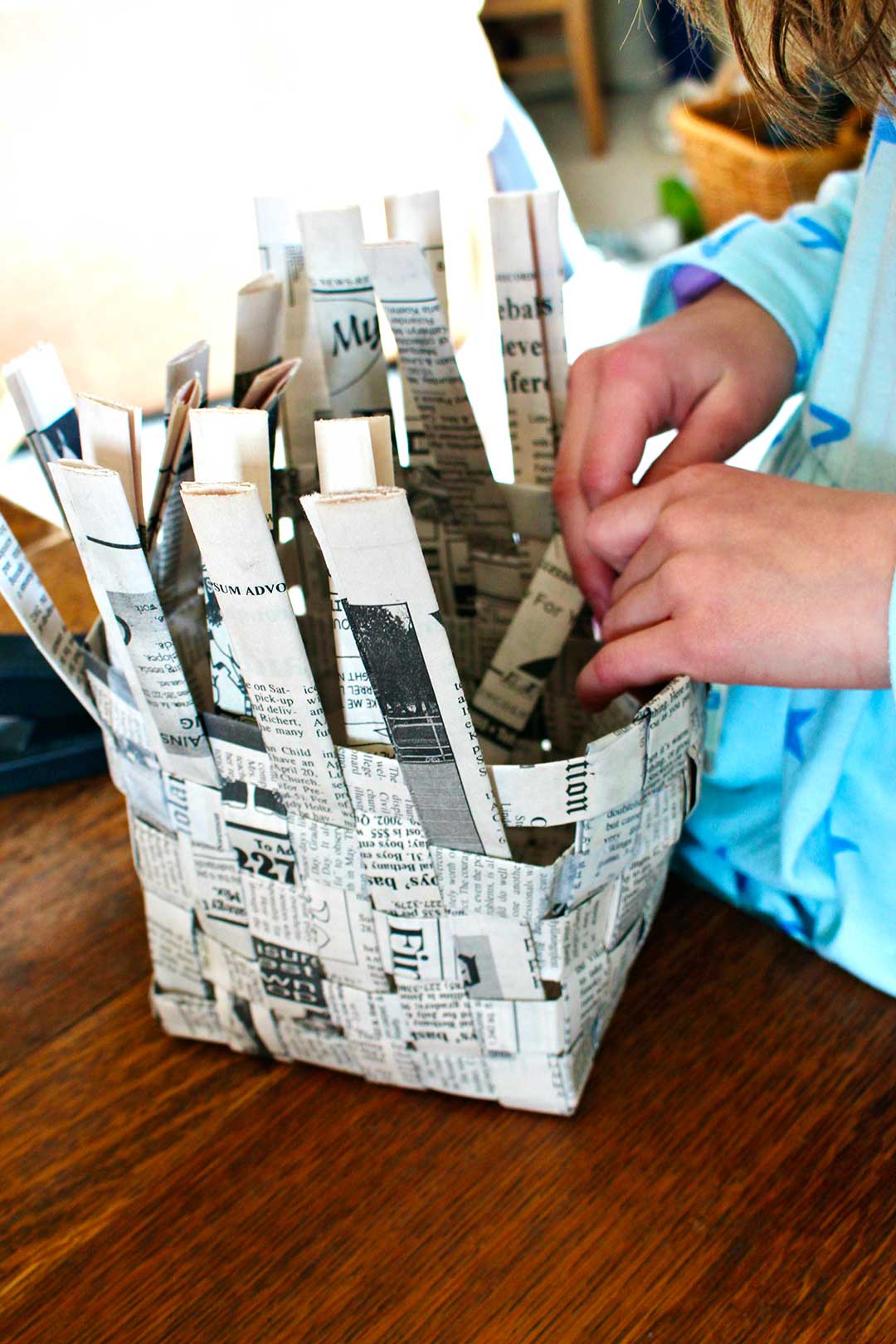 Child's hands folding down excess strips of newspaper to finish up woven newspaper basket.