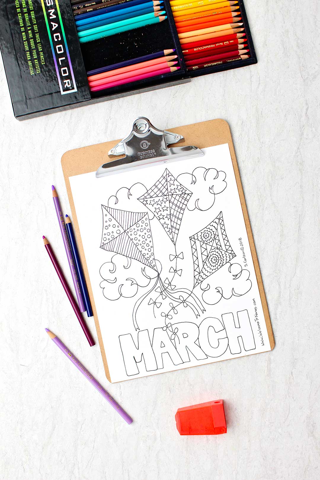 An uncolored March coloring page on a clip board with colored pencils near by. The page is a picture of three patterned kites in the sky with the word "March" at the bottom.