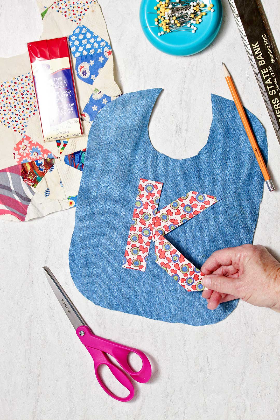 Hand placing a patterned piece of fabric to make the letter "K" on a blue bib base with supplies near by.