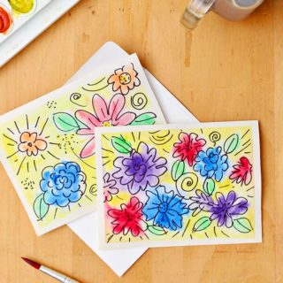 Two completed watercolor flower cards sitting on a light wood table with watercolors, marker, paintbrush and cup of water near by.