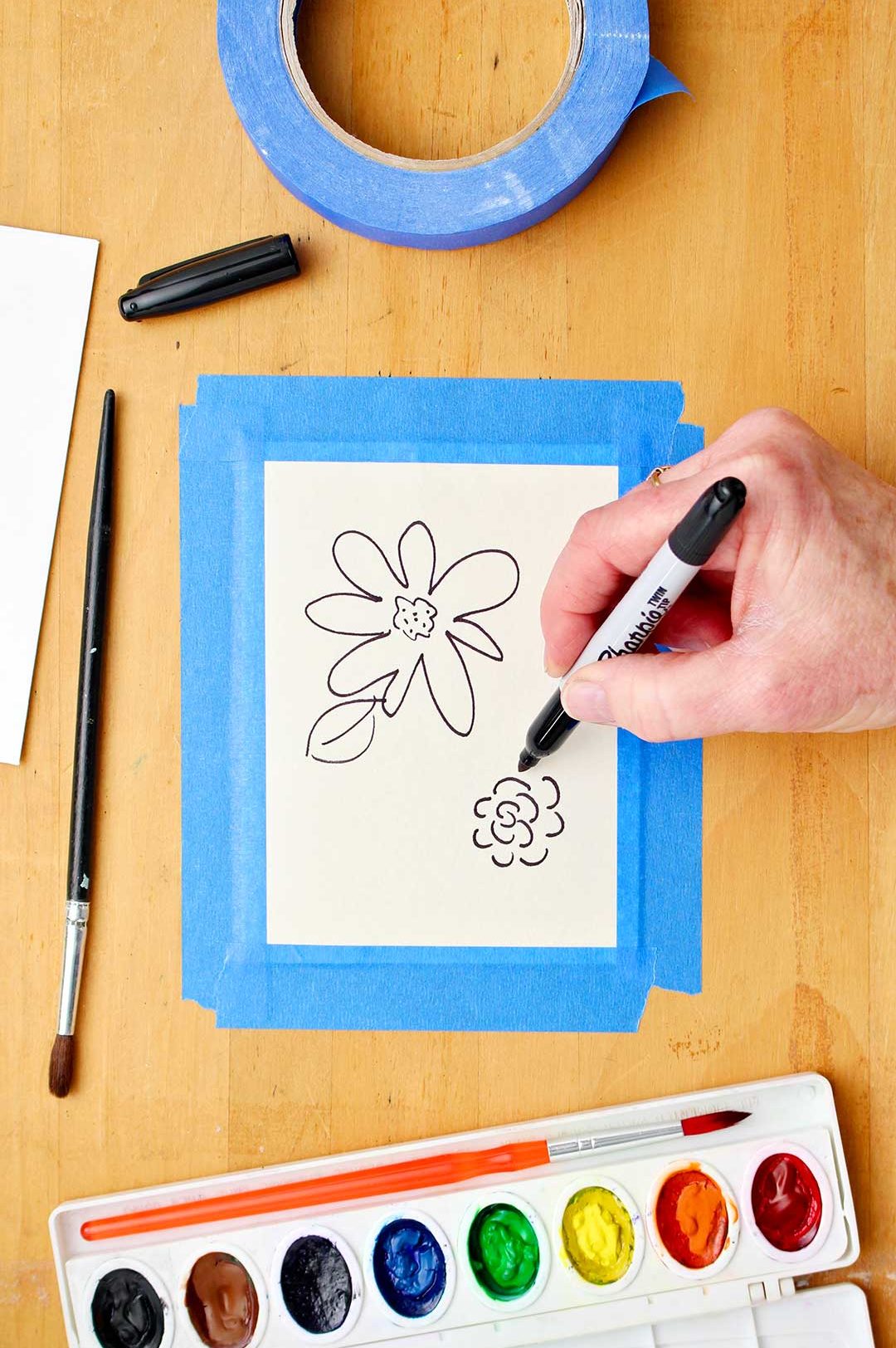 Hand drawing flowers with black Sharpie marker onto white card stock taped to a wooden table.
