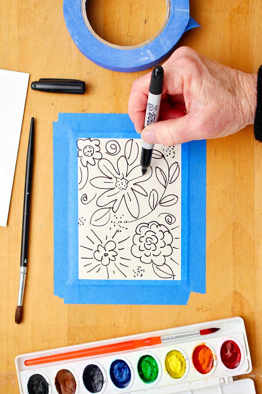 Hand drawing flowers and leaves with a black Sharpie marker on a wooden table with watercolor supplies near by.