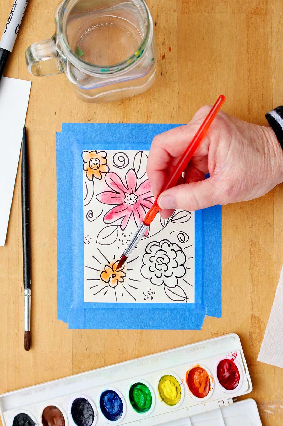 Hand painting marker outlined flowers orange with watercolors with supplies near by on table.